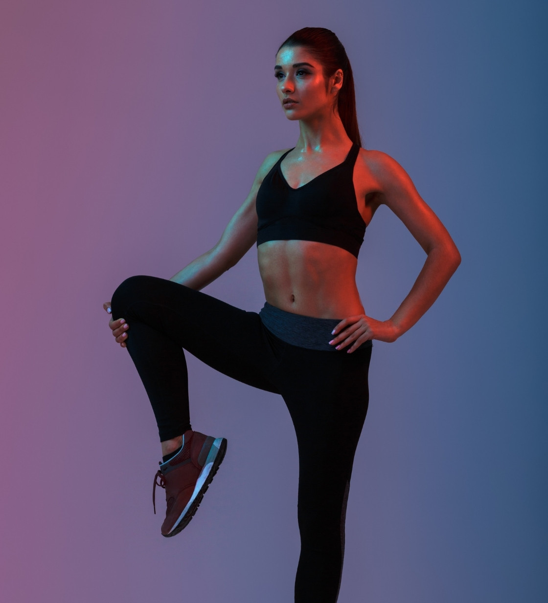 Full length portrait of concentrated young woman wearing black tracksuit doing sports and stretching her legs isolated over neon lights.